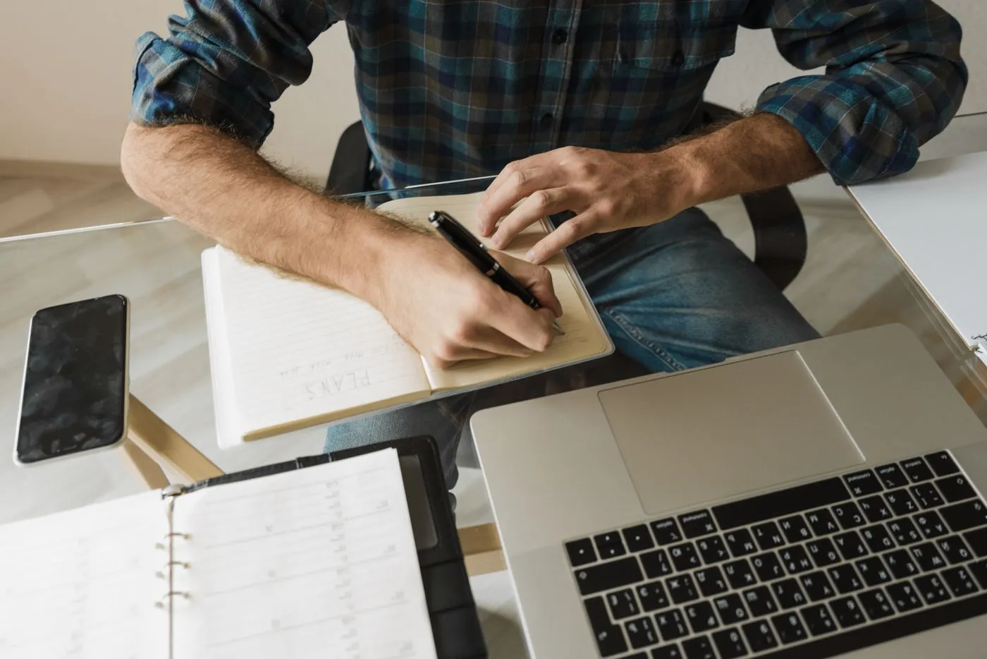 Hands writing a list in a notebook beside an open laptop